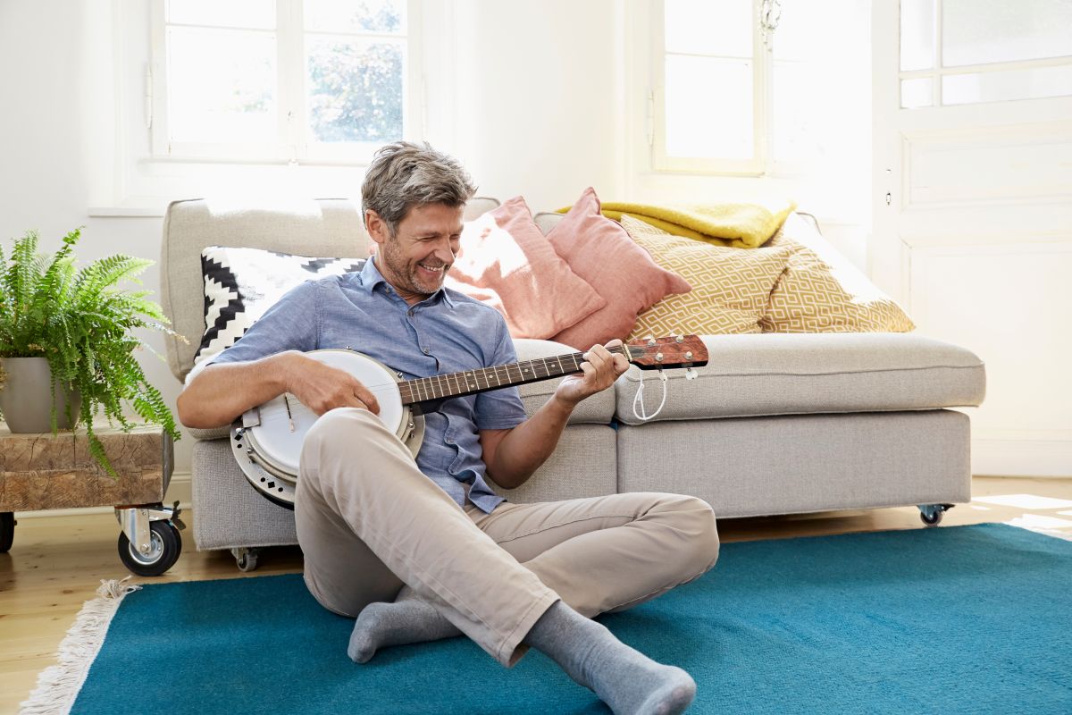 A man smiling while playing the banjo at home on the floor leaning against his couch